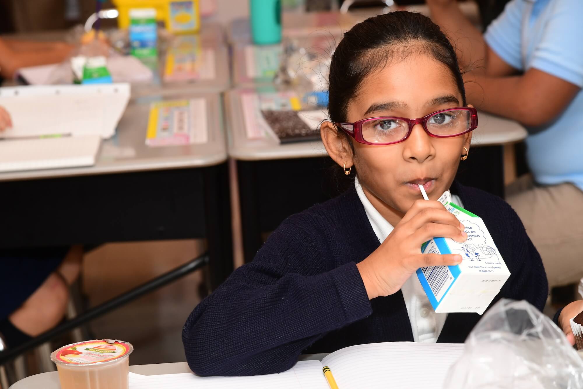 A girl wearing glasses drinks a carton of milk as part of her school lunch