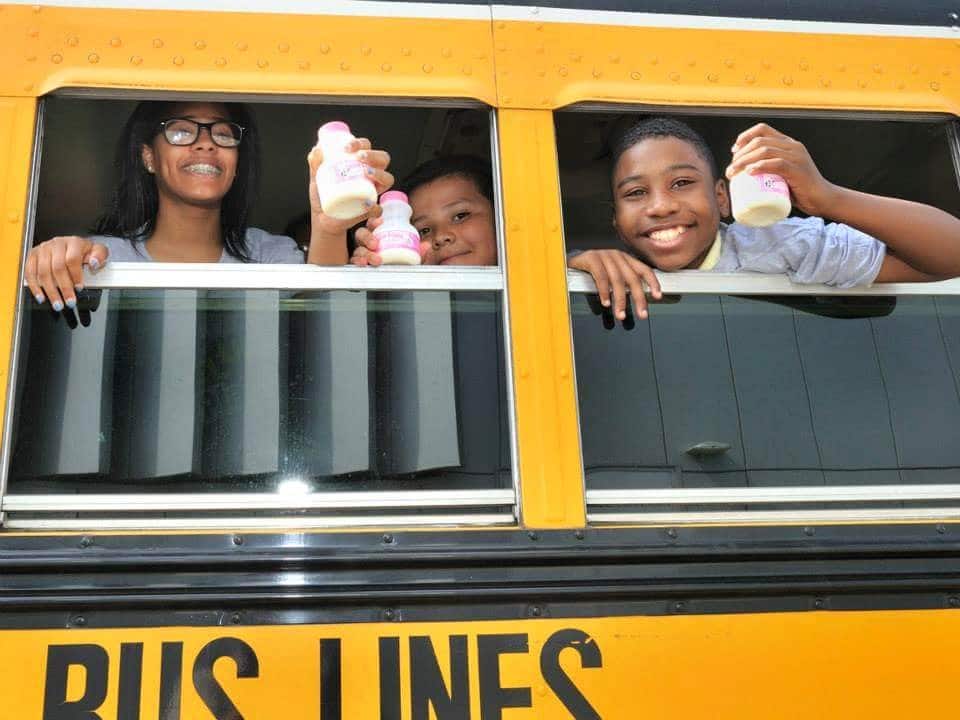 Children smiling and holding a bottle of milk each while peeking out a school bus window.